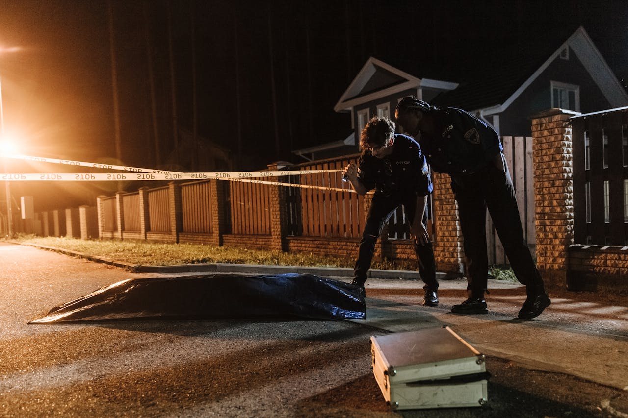 Police officers investigate a crime scene at night, examining evidence near a residential area.